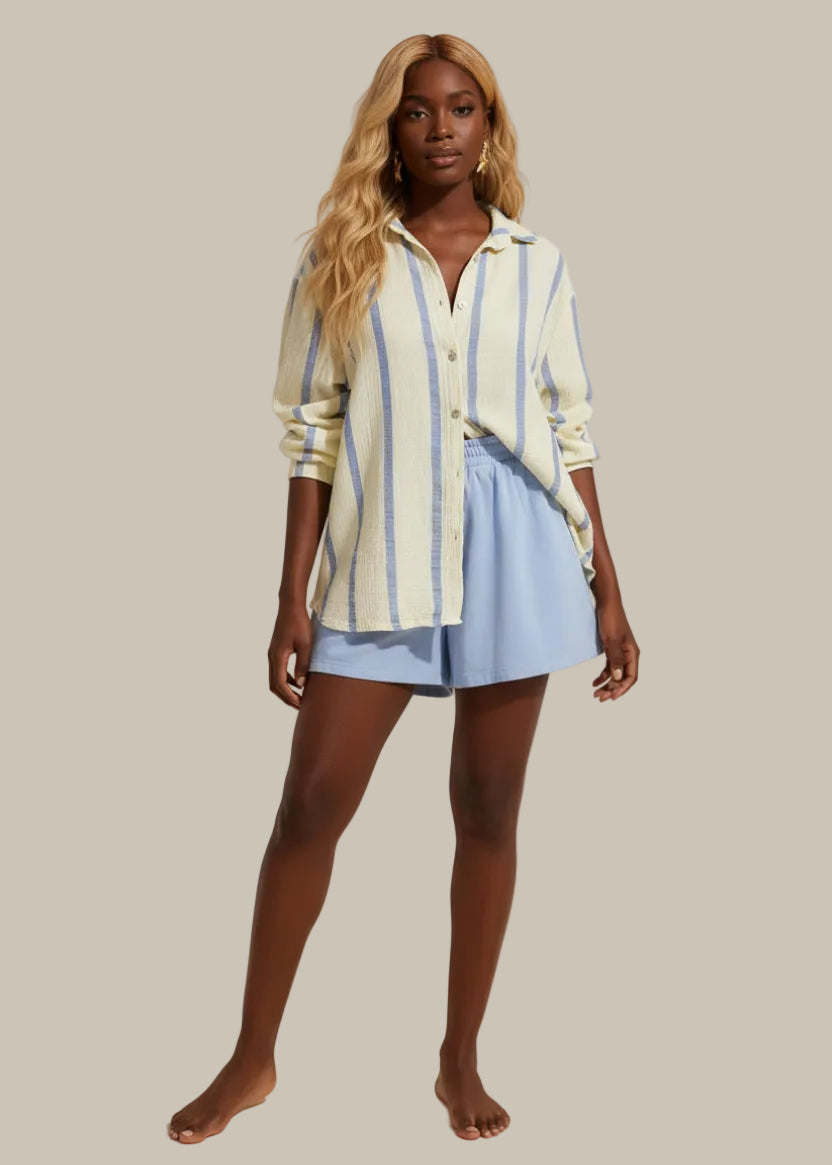 Woman wearing a striped shirt and blue shorts standing against a white wall.