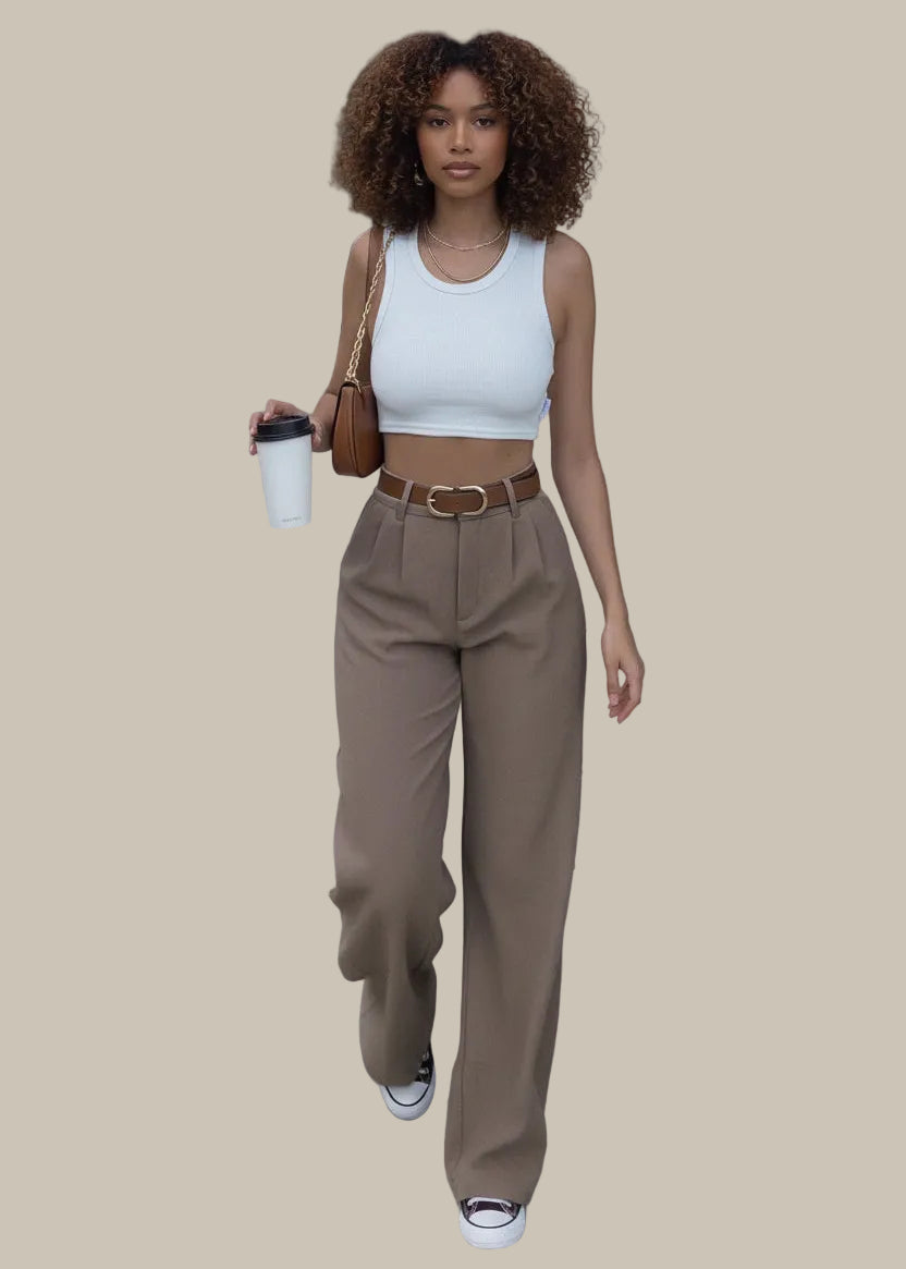 Person wearing a white crop top and brown high-waisted pants, holding a coffee cup on a sidewalk.