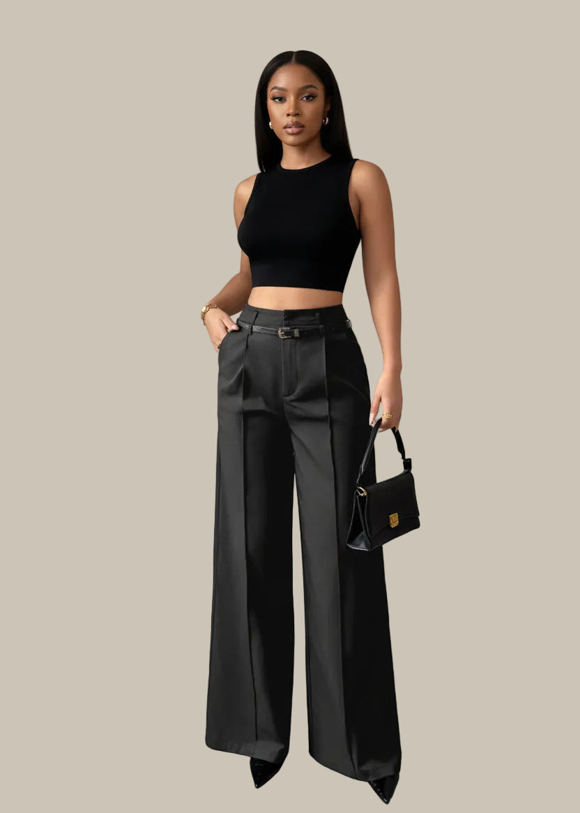 Person wearing black wide-leg pants and a black top, holding a black handbag against a gray curtain background.